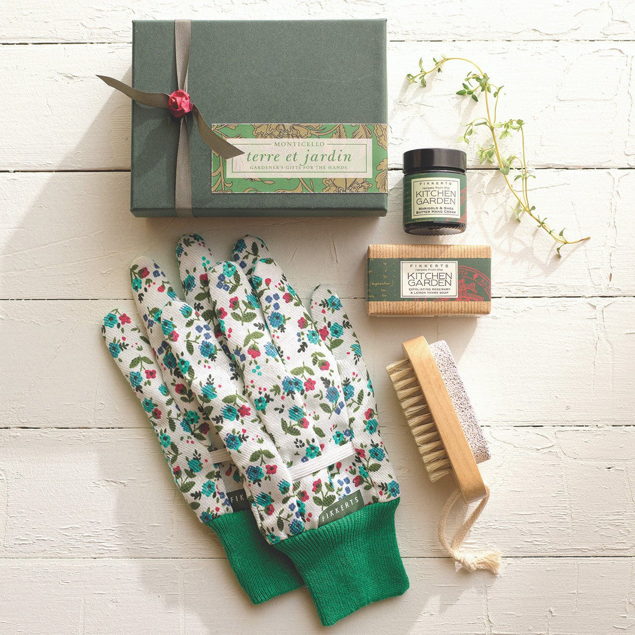 Gardening-themed gift set includes 3oz rosemary and lemon thyme bar soap, 1oz marigold & shea butter hand cream, nailbrush, pumice stone, and garden gloves, which are displayed on a coarse table